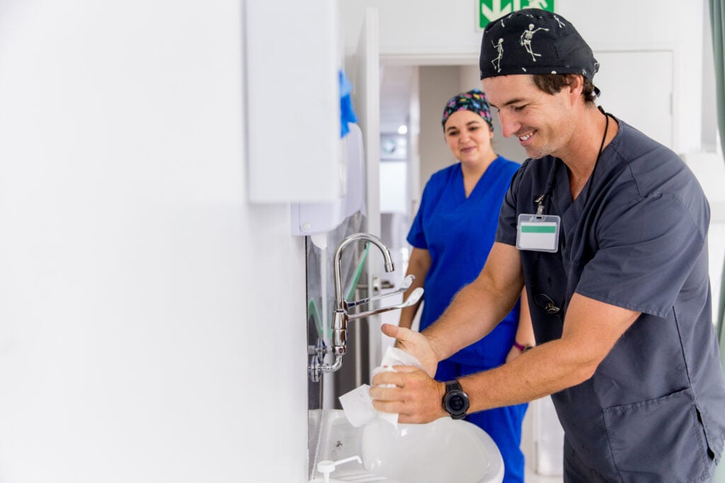 Care provider washing their hands in a hospital bathroom while another provider performs direct observation.